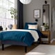 Modern bedroom: blue bed, grey walls, wooden floor. Mirror, plant, bedside table with lamp, clock. Shoes by bed. Window with curtains.