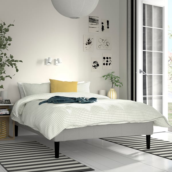 Modern bedroom with RENFJÄLLET bed, chequered duvet, gold pillow, striped rug, plants, art, lamps and window blinds.