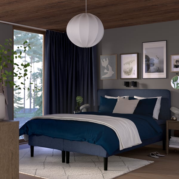 Minimalist bedroom: blue bed, dark walls, large window, pendant lamp, art, plants.