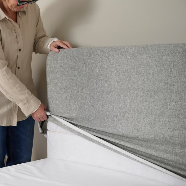 Person placing a grey bed headboard against a wall. The headboard appears comfortable and designed to lean against without disturbing sleep.