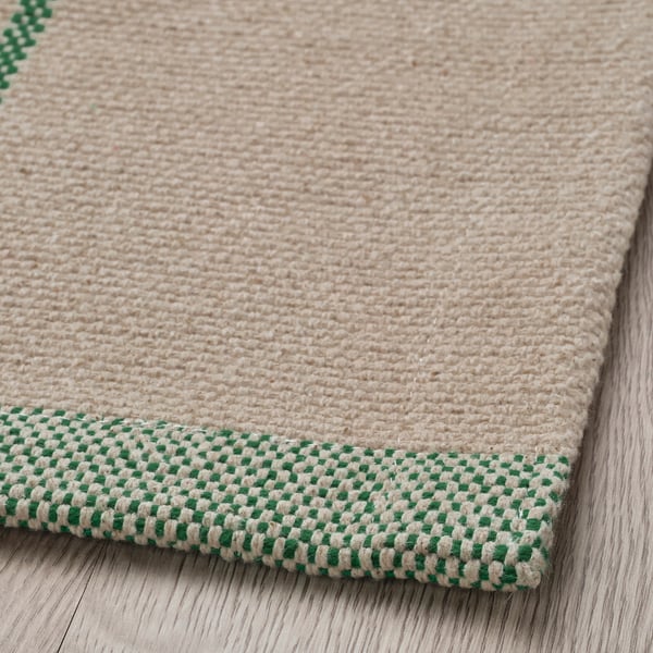 Beige woven rug with green checkered border, placed on a wooden floor.