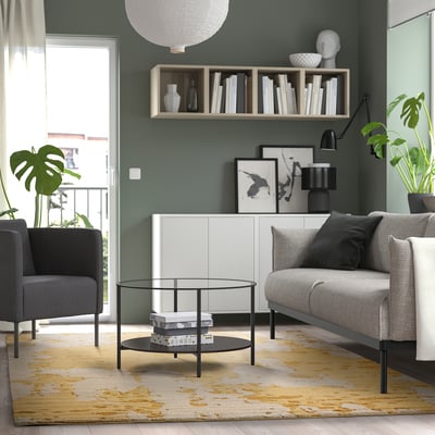 Modern living room with yellow & beige rug, sleek furniture, plants, & large windows.