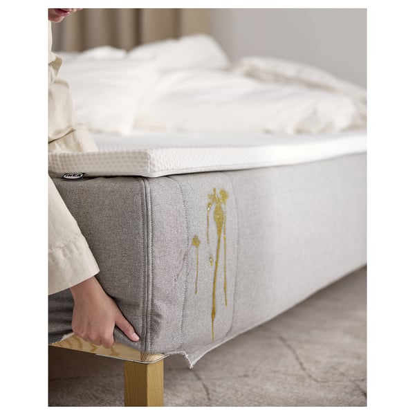 A person adjusting a RISHÖJDEN bed, white and grey with gold accents, made from recycled polyester and polypropylene, on a wooden base.