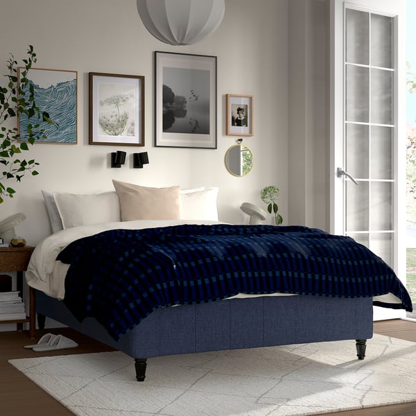 Modern bedroom with RISHÖJDEN bed, navy blue plaid, wooden frame, art wall, cushy headboard, white & beige pillows, side lamp, green plants.
