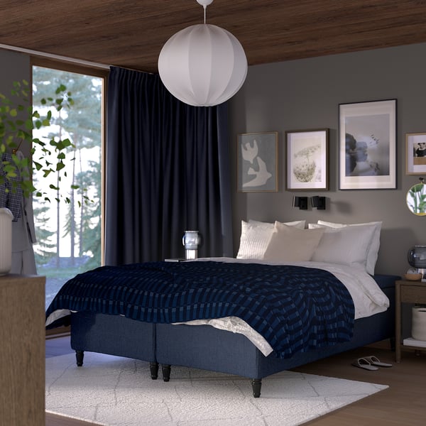 Dark blue bed with white pillows, large window, grey walls, wooden ceiling, hanging lamp.
