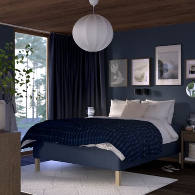 Modern bedroom with dark blue walls, blue bed, white linens, pendant light, wooden side table, lamp, plant.