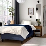 Modern bedroom: blue bed, white pillows, wooden bedside table with lamp, clock, and plant. Large window with city view.