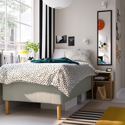 Modern bed RISHÖJDEN with polka dot bedding by large window, beside wooden table with books and bag.