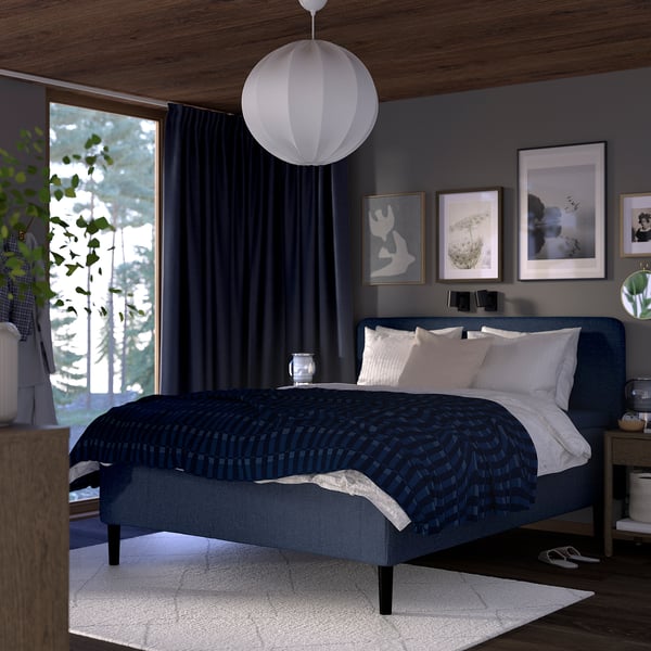 Modern blue RISHÖJDEN bed with soft headboard, large window, white lampshade, art, dark curtains, wooden floor, white rug.