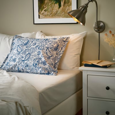 Bedroom with RODGERSIA paisley pillowcases, beige bedding, books, dried flowers, warm lamp.