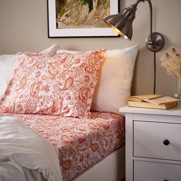 Cozy bedroom scene with RODGERSIA coral paisley pillowcase on a bed, white pillows, a book, lamp, and vase on a nightstand.