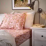 Cozy bedroom scene with RODGERSIA coral paisley pillowcase on a bed, white pillows, a book, lamp, and vase on a nightstand.