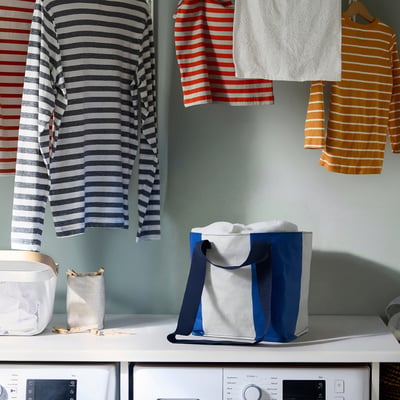 Striped shirts hang near dryer; a white, blue, and grey modern SÄCKKÄRRA bag sits on top, small, compact, and trendy.