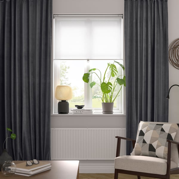 Modern living room with large SANELA dark grey curtains framing a tall window, featuring a plant, lamp, and cozy chair with a patterned pillow below.