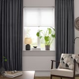 Modern living room with large SANELA dark grey curtains framing a tall window, featuring a plant, lamp, and cozy chair with a patterned pillow below.