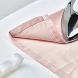 Image displays a pair of pale pink SANELA curtains with a silver iron resting on them. Curtains are neatly folded with a visible seam.