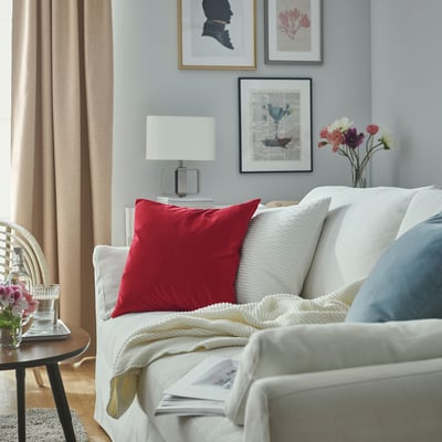 Modern living room with white sofa, red SANELA cushion, table lamp, and flower vase on wooden table, blue curtains.