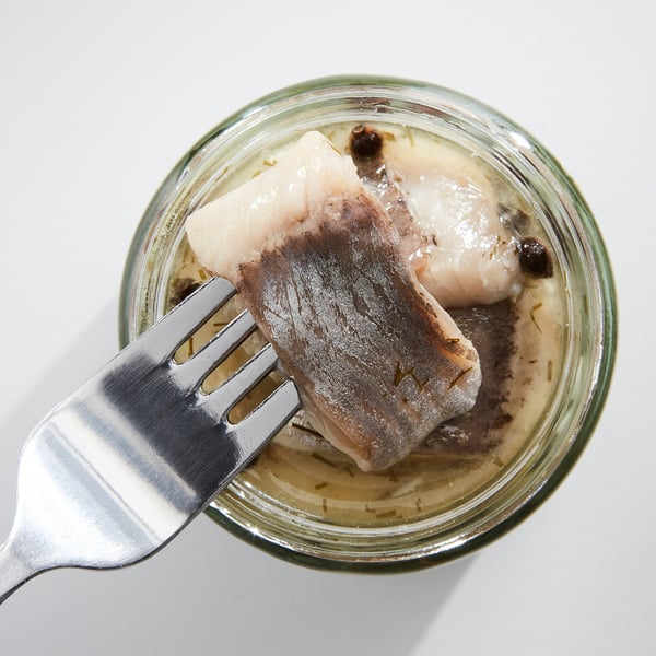 Fork lifting pickled fish from glass jar on white table.