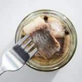 Fork lifting pickled fish from glass jar on white table.
