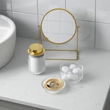 Bathroom vanity set with gold accents: circular mirror, glass jar with lid, and bowls for cotton and jewelry.