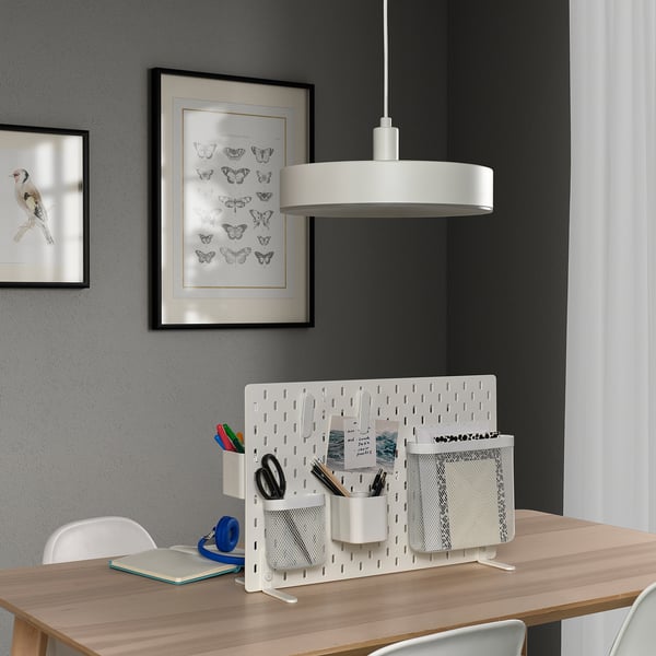 SKÅDIS white desk divider with pockets on wooden desk. Pens, scissors, notebook. Two wall art pieces. Central light.