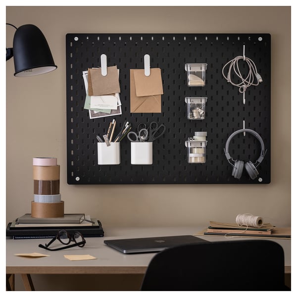 SKÅDIS black pegboard organises office supplies with accessories: clips, cups, and hooks on beige wall.