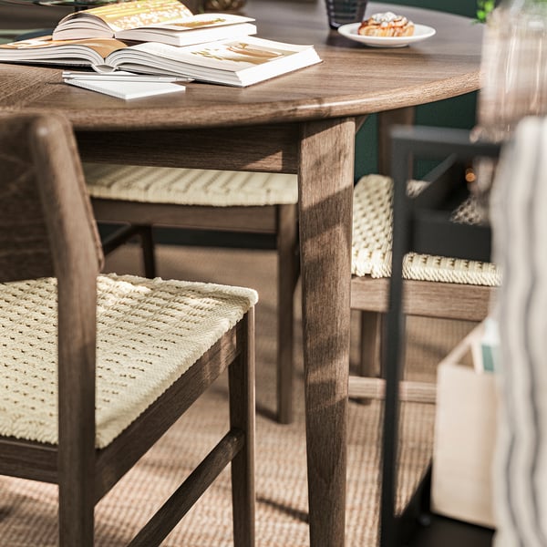 SKANSNÄS round dining table in brown; wooden texture, seats six. Books stacked and plate beside. Woven chairs complete set; shown near rug.