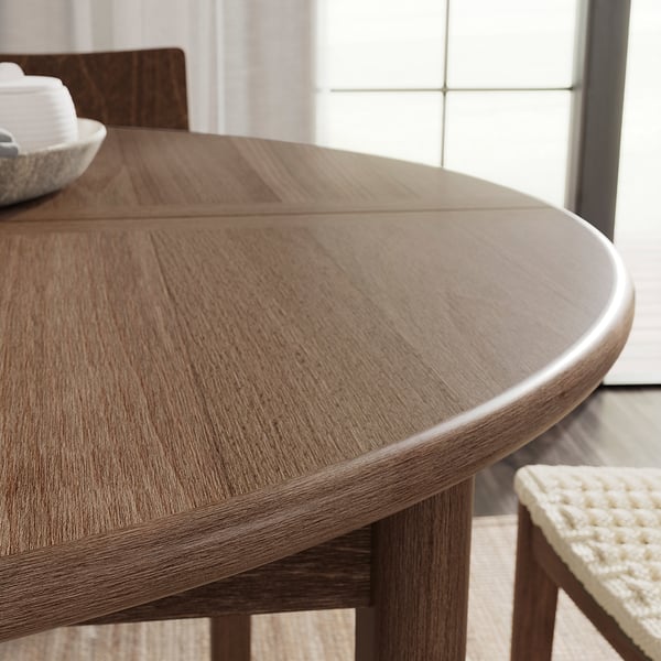 Brown wood round dining table with smooth surface and sleek design, positioned near a window.