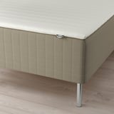 A neatly made bed with a white mattress and beige upholstered base, elevated by metal legs from SKÅRER.