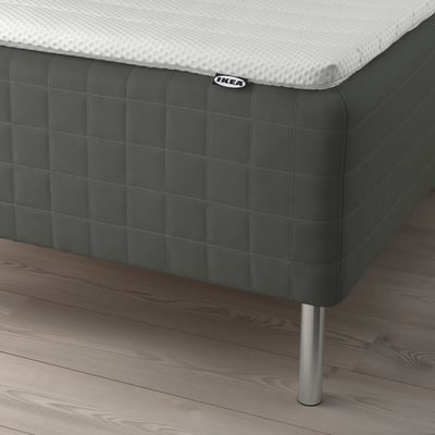 A bed with a gray SKOTTERUD sprung mattress on a dark floor. The mattress has a white top and is supported by metal legs.