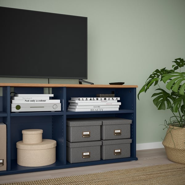 A blue tv stand with open shelves, storing books, boxes, and electronics. A large flat-screen tv is placed on top. A potted plant is nearby.