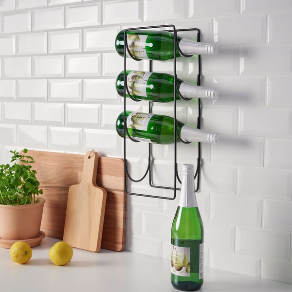 Wall-mounted metal wine rack holding four green bottles, two lemons, wooden cutting boards, and a potted plant on a white countertop.
