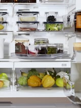 An organized refrigerator with white SNURRAD storage trays holding various fruits, vegetables, and glass containers of food.