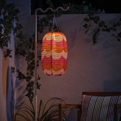 Lit lantern hangs from white stand, casting warm colours against a textured wall, surrounded by green plants.