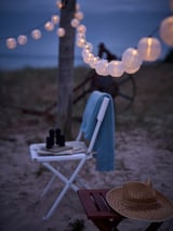 Cozy beach setup at dusk with string lights, white folding chair, hat, and table.