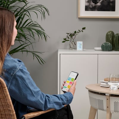 A woman adjusts a smart air purifier via app. The device monitors air quality and can be controlled remotely. It sits on a cabinet with decor items.