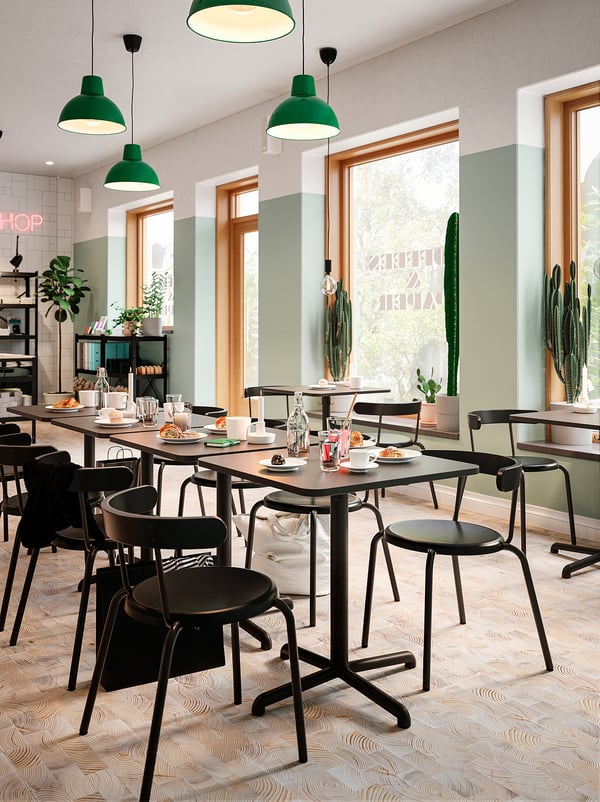 Modern café with square STENSELE tables in black, green pendant lights, large windows, and cacti.
