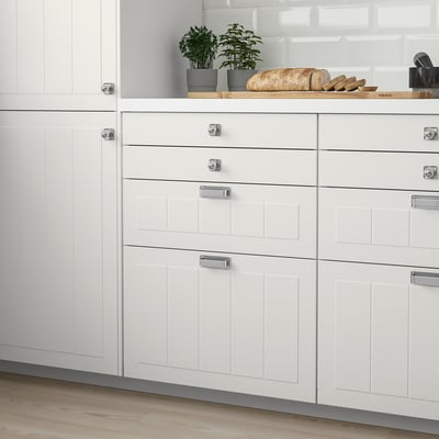 Modern white kitchen cabinets with metal handles, greenery, and light floors.
