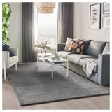 Stylish living room with grey STOENSE rug, grey sofa, colorful pillows, white coffee table, large window, grey curtains, and plant.