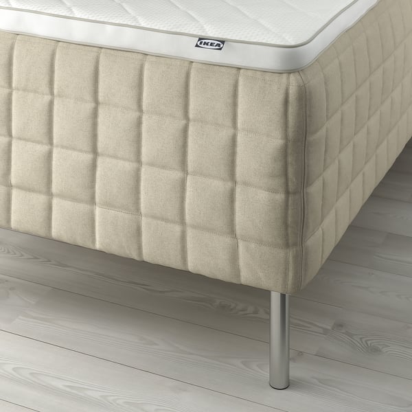 Close-up of beige IKEA mattress on wooden floor. Shows quilted side with subtle texture, metal legs, and white branding.