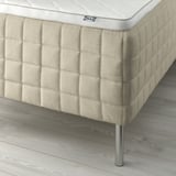 Close-up of beige IKEA mattress on wooden floor. Shows quilted side with subtle texture, metal legs, and white branding.