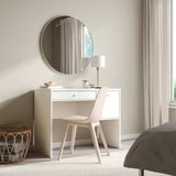 Minimalist bedroom with white SYVDE vanity. Round mirror, lamp, wicker basket. Clean, simple design. Soft-close drawers. Beige carpet.