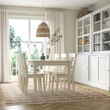 Bright dining room with white furniture, warm rug, woven light, plant, and large window.