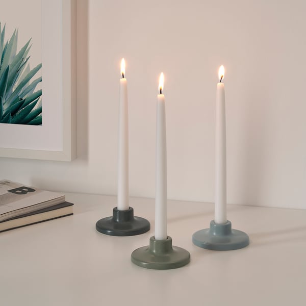 Three lit candles in colorful holders, arranged on a white surface with a book and plant illustration nearby.