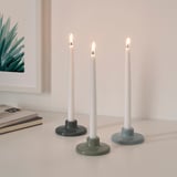 Three lit candles in colorful holders, arranged on a white surface with a book and plant illustration nearby.