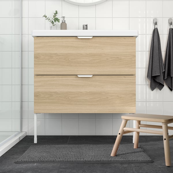 Modern bathroom with light wooden vanity, black bath mat, and wooden stool.