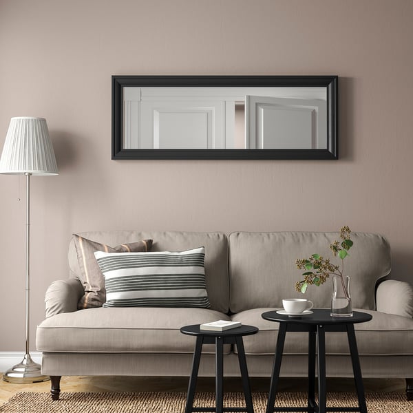 Black TOFTBYN mirror hangs above beige sofa, reflection shows doorway, flanked by black side tables with flowers and tea set.