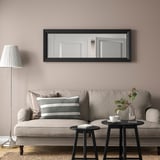 Black TOFTBYN mirror hangs above beige sofa, reflection shows doorway, flanked by black side tables with flowers and tea set.