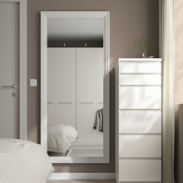 Minimalist bedroom with large TOFTBYN mirror reflecting white wardrobe, beside narrow white drawer unit with plant.
