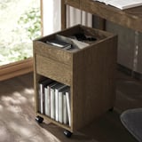 Mobile wooden storage unit with drawer and shelf, holding books and office items. Ideal for small spaces.